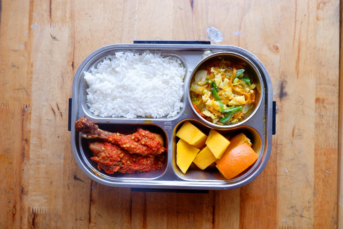 A partitioned lunchbox filled with rice, chicken, curry, and fruit.
